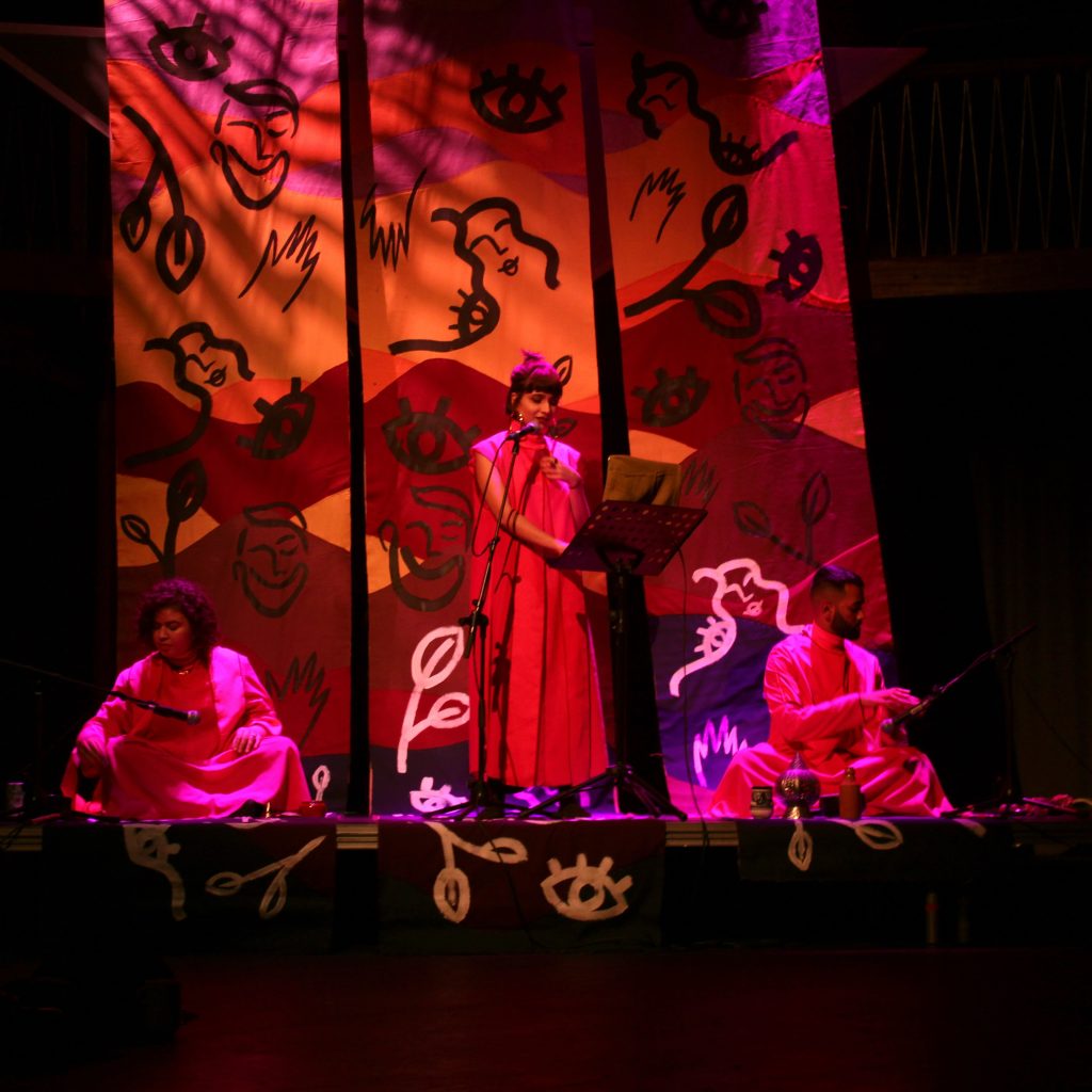 Woman in red speaks into a microphone on stage, in front of an orange background.