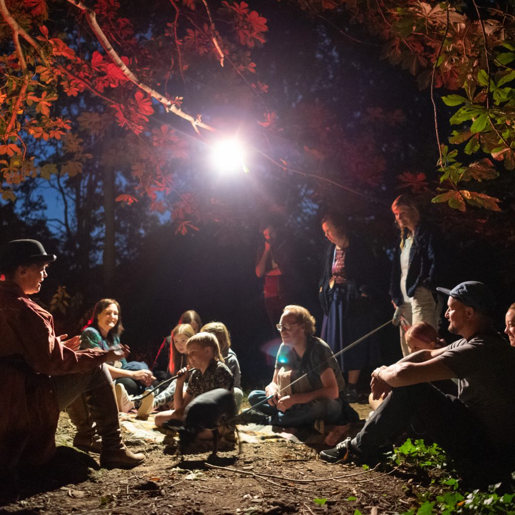 Children sat in a circle on the ground outside in a forest/woodland area while listening to storues