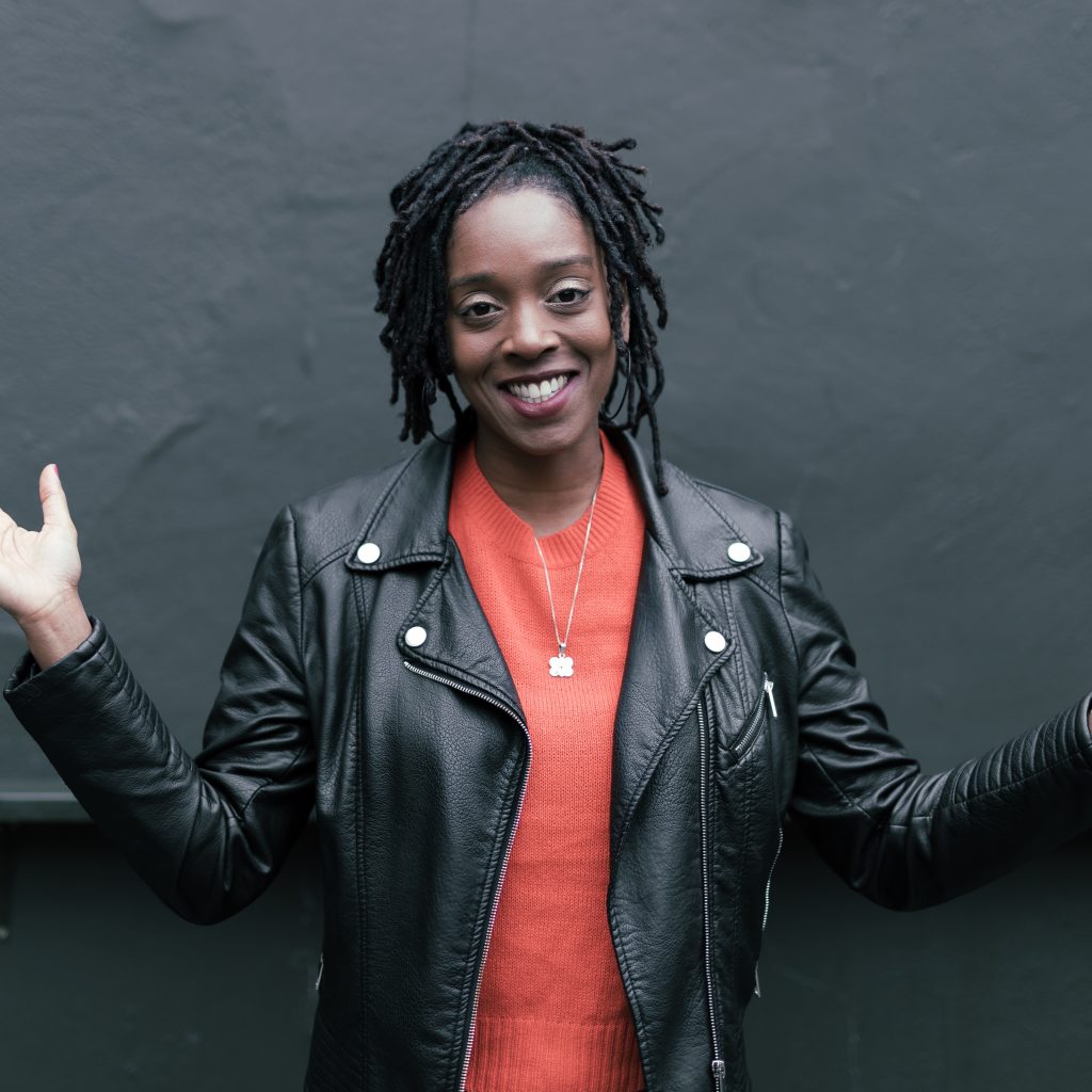 Photo of Athena Kugblenu wearing a red shirt and leather jacket, smiling at the camera